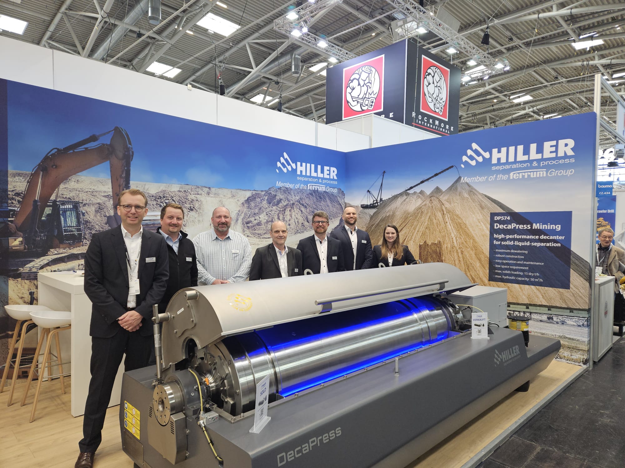 Seven people in business attire stand beside a large industrial DecaPress machine at the HILLER at Bauma 2025 exhibition booth, with mining and machinery banners displayed in the background.