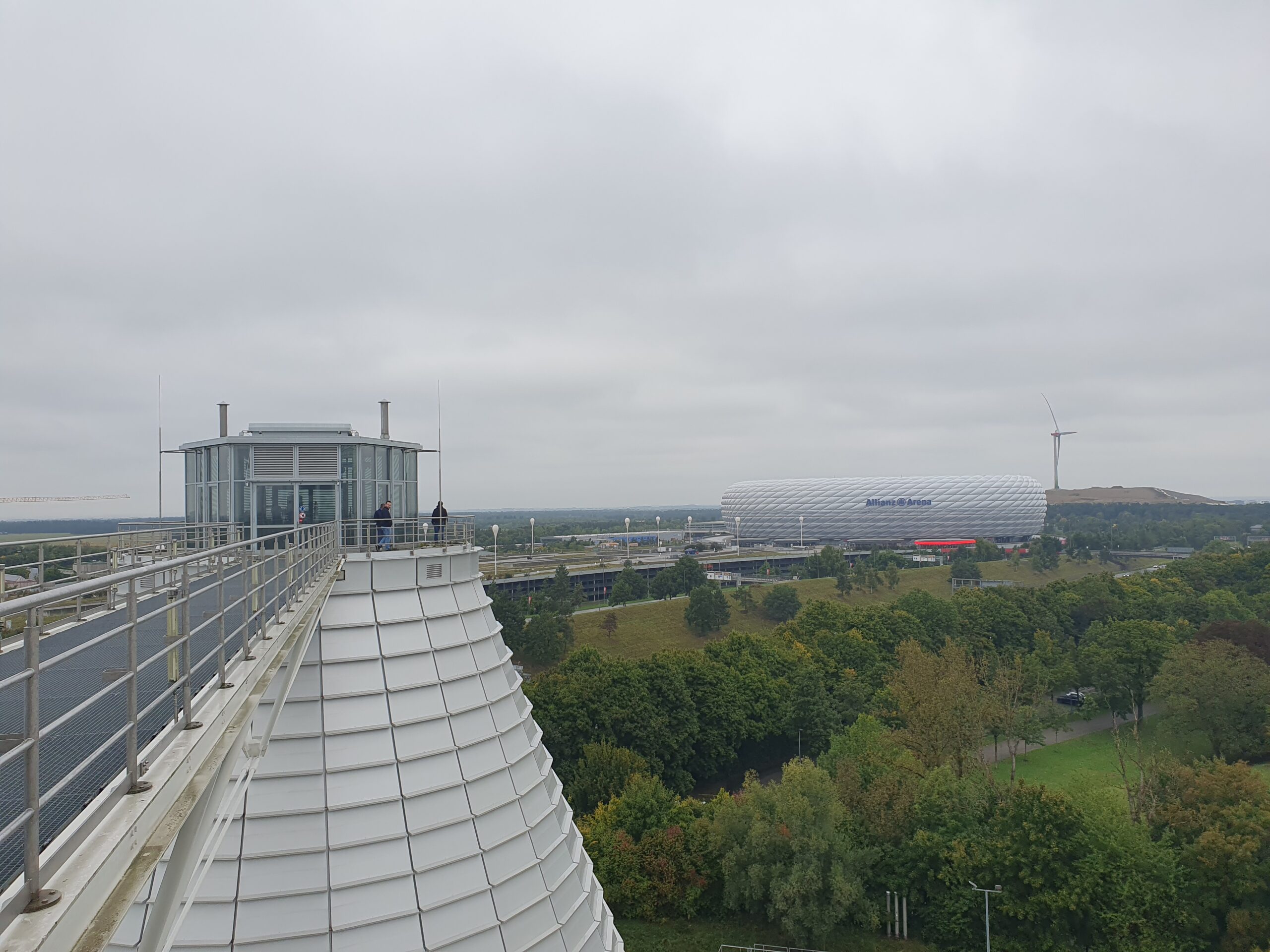 View from a rooftop platform overlooking a modern, white-domed stadium, surrounded by greenery and trees under a cloudy sky. A wind turbine and distant hills are visible in the background.