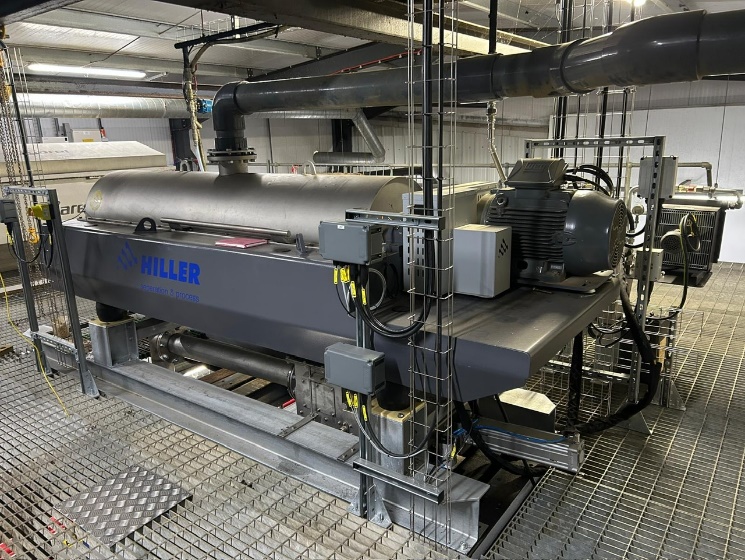 A large industrial machine, labeled Hiller, sits on a metal grated floor inside a factory. Pipes and cables are attached to the machine, and industrial lighting illuminates the workspace.