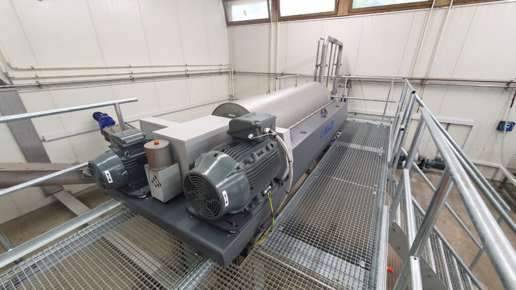 Industrielle Zentrifugenmaschine mit zwei großen Motoren, installiert auf einer Metallgitterplattform in einer sauberen, weißwandigen Anlage mit hohen Fenstern und sichtbaren Rohrleitungen.