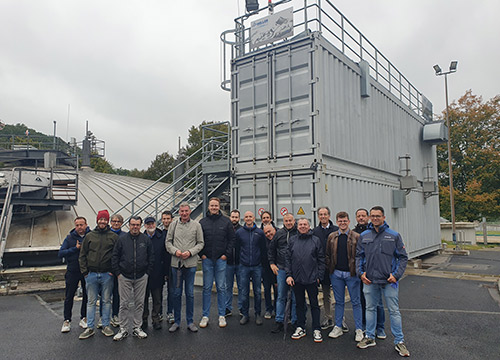 Eine Gruppe von Menschen in Jacken und Jeans steht an einem bewölkten Tag im Freien vor einem großen, grauen Industriecontainer mit Treppe und Geländer zusammen. Im Hintergrund sind Bäume und andere industrielle Strukturen zu sehen.