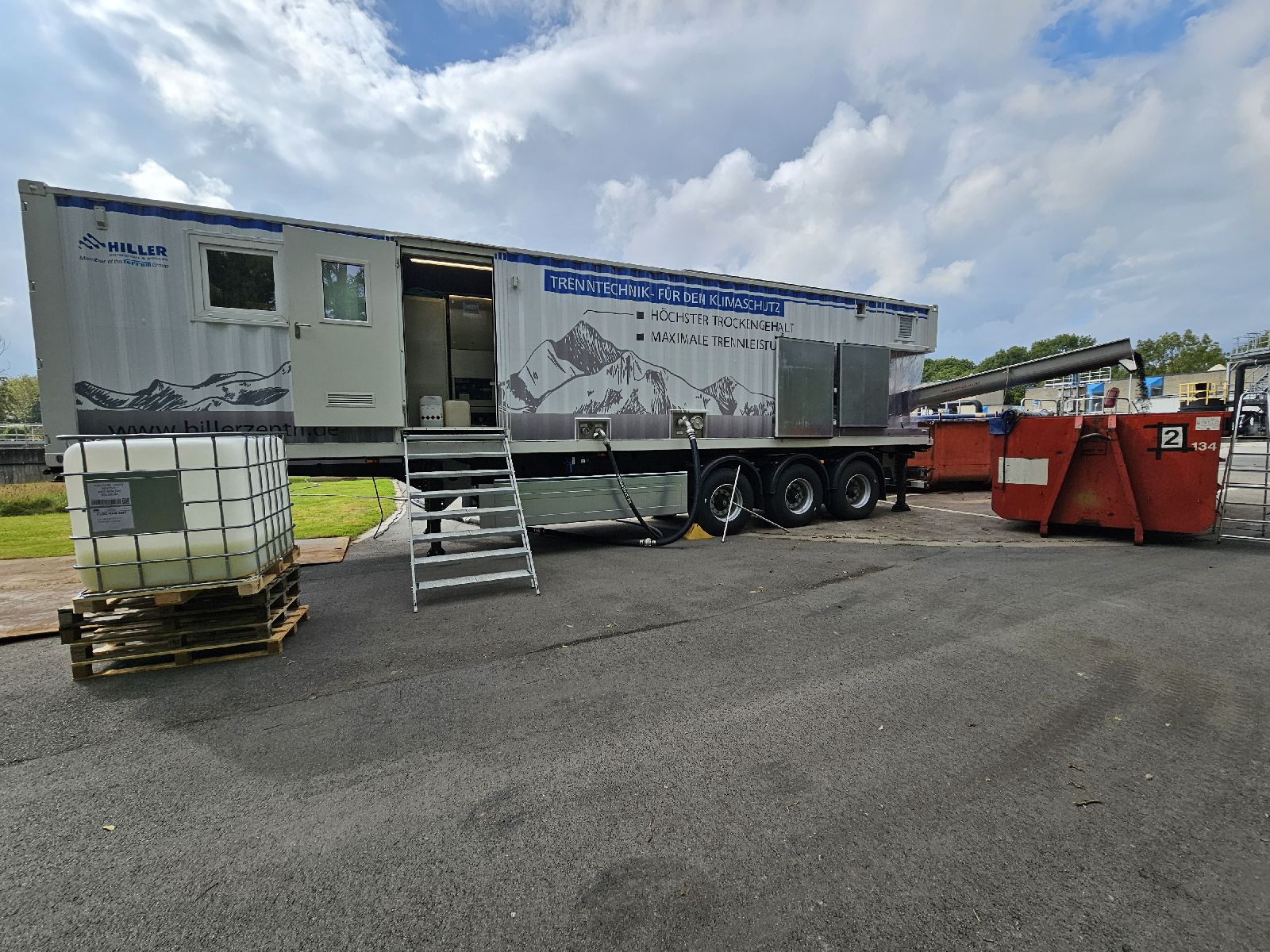 A large mobile unit with “Trenntechnik für den Kanalspül” written on its side is parked on asphalt. Steps lead to the entrance. Large tanks and red waste containers are nearby. The sky is partly cloudy.