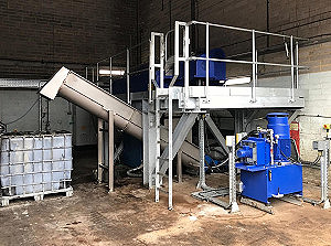 A metal industrial machine with a conveyor belt and blue motor components stands in a warehouse. Nearby is a large white plastic container with a metal frame on a stained concrete floor.
