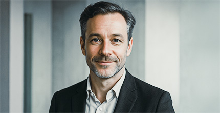 Ein Mann mit kurzen dunklen Haaren und gestutztem Bart, der ein weißes Hemd und einen dunklen Blazer trägt, lächelt sanft, während er in einer modernen, hellen Büroumgebung steht.