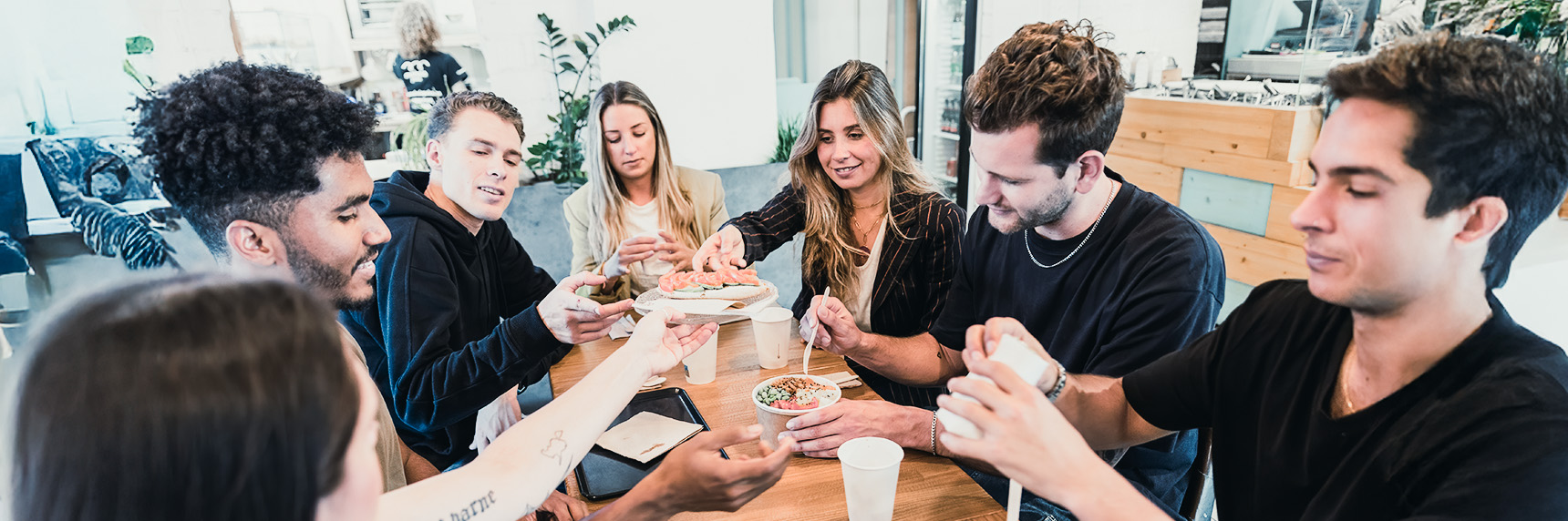 Eine Gruppe von sechs jungen Erwachsenen sitzt um einen Tisch herum, lächelt und isst gemeinsam in einer ungezwungenen, modernen Innenumgebung. Sie scheinen die Mahlzeit und die Gesellschaft der anderen zu genießen.