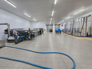 Large, clean industrial facility with stainless steel tanks on the right, machinery and two people working on the left, and blue hoses lying on the tiled floor. A car and more equipment are in the background.