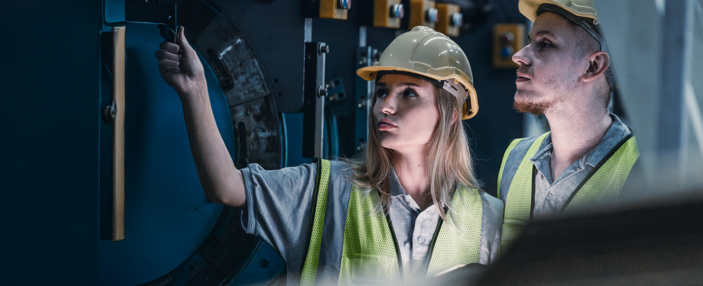 Zwei Arbeiter, die gelbe Schutzhelme und Warnwesten tragen, untersuchen Maschinen in einer industriellen Umgebung, wobei eine Person auf die Bedienelemente zeigt, während die andere aufmerksam beobachtet.