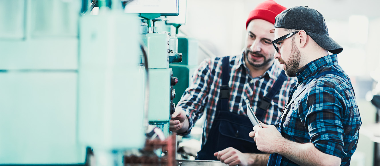 Zwei Männer in karierten Hemden und Hüten arbeiten gemeinsam an einer Maschine in einer Industrieumgebung. Der eine zeigt auf die Bedienelemente, während der andere ein Tablet in der Hand hält, beide konzentriert auf ihre Aufgabe.