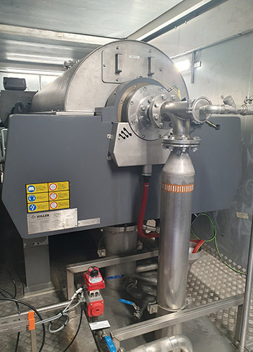 Industriemaschine mit einem großen Metallfass und angeschlossenen Rohren, Ventilen und Schalttafeln in einer Anlage. Warn- und Hinweisschilder sind an der Seite des Geräts sichtbar.