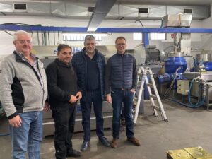 Four men stand side by side in an industrial setting with machinery, pipes, and equipment in the background. They are dressed in casual and semi-casual clothes, posing and smiling for the photo.