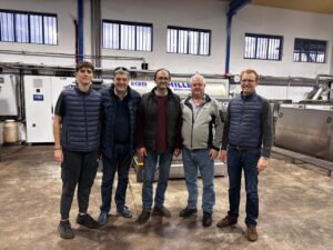 Five men wearing jackets and casual clothing stand indoors on a polished concrete floor in front of industrial equipment and large windows, posing and smiling for a group photo.