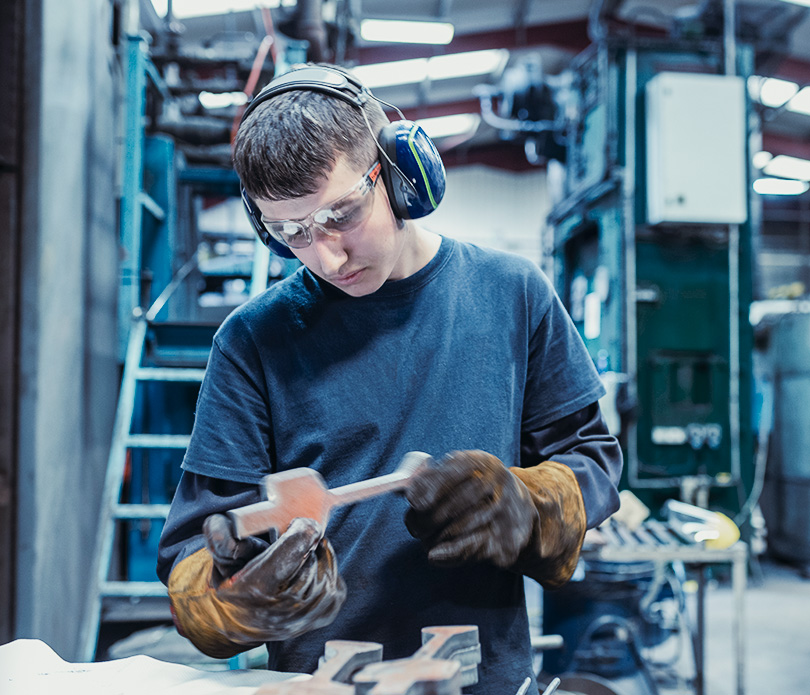 Ein junger Mann mit Schutzbrille, Gehörschutz, Handschuhen und dunklem Hemd prüft ein Metallwerkzeug in einer industriellen Werkstatt. Im Hintergrund sind Maschinen und Geräte zu sehen.