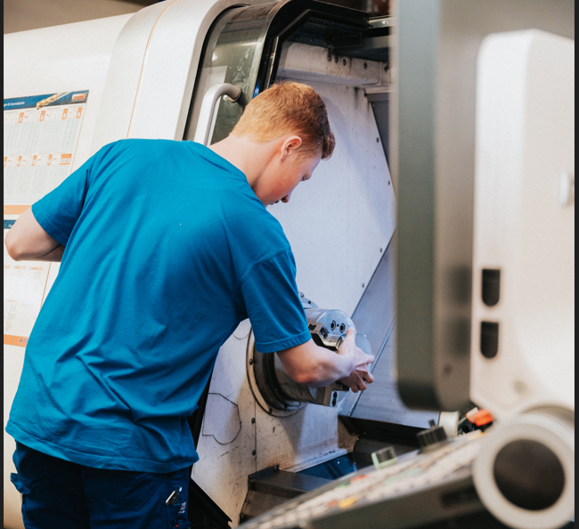 Eine Person in einem blauen Hemd bedient eine CNC-Maschine und greift in das offene Fach, um möglicherweise ein Bauteil einzustellen oder zu überprüfen, in einer Industrie- oder Werkstatteinrichtung.