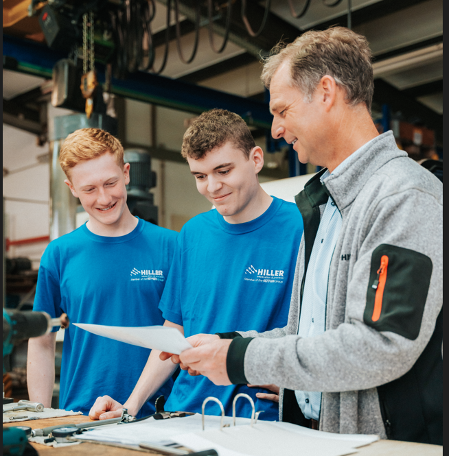 Ein Mann in einer grauen Jacke hält Papiere in der Hand und unterhält sich mit zwei lächelnden jungen Männern in blauen Hiller-T-Shirts in einer Werkstatt, in der im Hintergrund Werkzeuge und Geräte zu sehen sind.